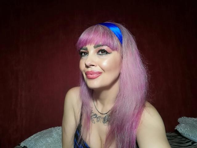 HotAdelayde69 a les cheveux roses avec un bandeau bleu, la langue tirée et un tatouage au-dessus de ses seins. Elle porte un haut bleu avec des bretelles noires..