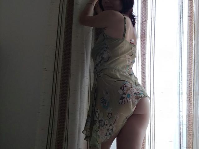 Cassy's back is to the camera, wearing a floral dress, showing her bare ass. She's standing by a window, her hand on the curtain, with pale skin.