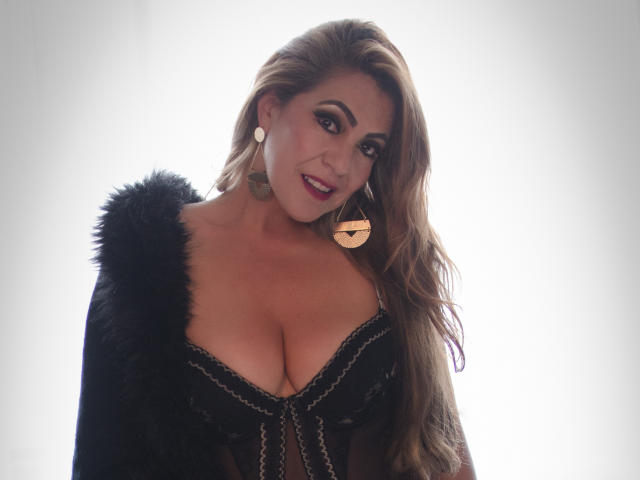 LylaGreen with long brown hair, wearing a black sheer dress with fur shoulders, exposing her large tits, and gold earrings, smiling at the camera.