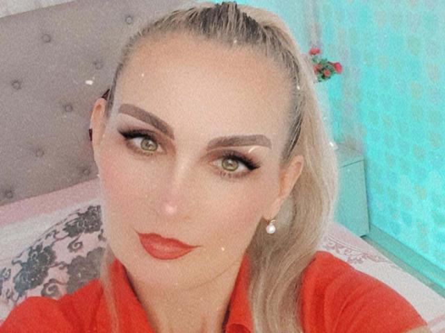 Una mujer atractiva con cabello rubio recogido en una coleta, lápiz labial rojo y ojos verdes, vestida con una camisa roja, aretes de perlas y delineador de ojos llamativo, sonriendo suavemente..