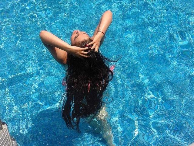 LexyRene with wet black hair, hands in hair, blue-tinted tits, floating in clear pool, arms raised, sunlight rippling water, looking up, pink swimsuit barely visible.