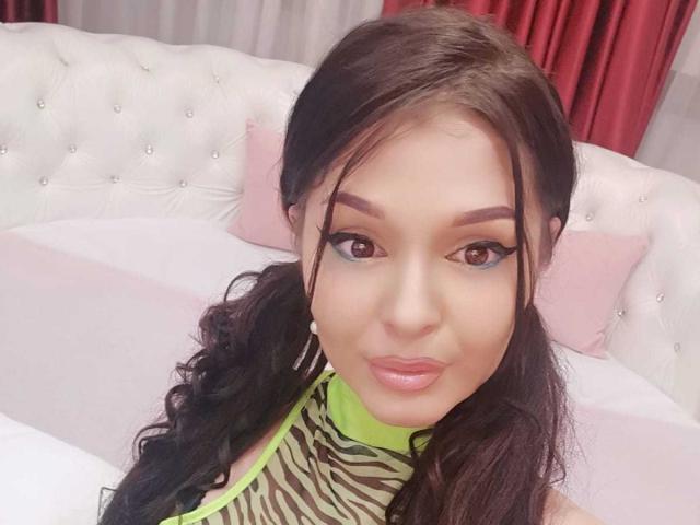 LexyRene, com lábios rosados e expressivos, olhos castanhos e cabelo castanho escuro preso em um rabo de cavalo, vestindo uma blusa listrada como zebra com uma gola verde neon. Uma cama branca com detalhes em relevo fica atrás..