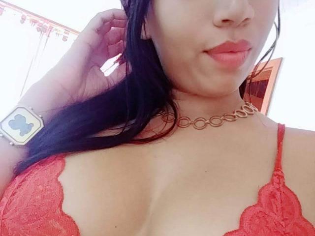 AlanaMorgan69 draagt een rode kant BH, waardoor haar grote borsten zichtbaar zijn, een bruine choker, zwart haar en roze lippenstift. Ze draagt een horloge met een blauw kruis erop..