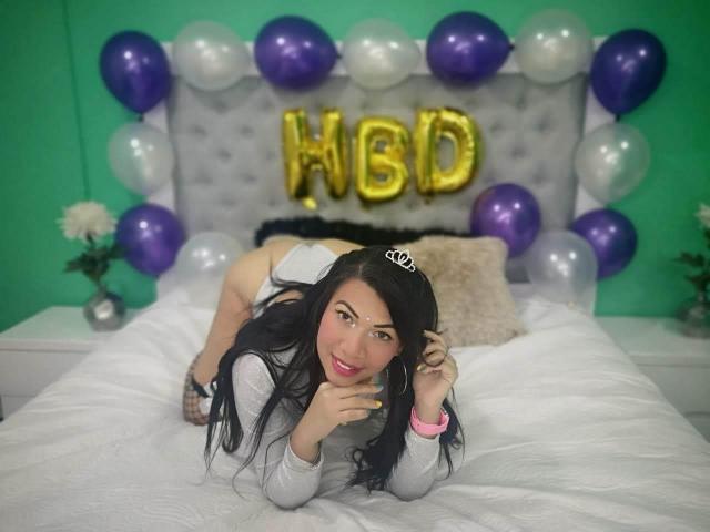 AlanaMorgan69 ligt op een bed, gekleed in een witte jurk en een kroon, met paarse en witte ballonnen en een achtergrond van ballonnen met de letters "WBD". Haar billen zijn zichtbaar, en ze kijkt naar de camera..