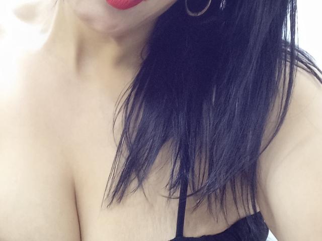 LizaRussell's black hair, red lipstick, and bare cleavage with a black strap showing her tits. She's wearing gold hoop earrings. Soft skin, slight tan. Upper body shot.