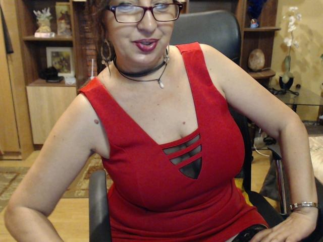 LadyEvelynn trägt ein rotes Kleid, auf dem ihre Brüste sichtbar sind, sitzt auf einem Stuhl, trägt eine Brille, eine schwarze Halskette und ein Armband, lächelt und befindet sich im Innenbereich. Ihre Brüste haben ein kleines Tattoo auf der rechten Seite..