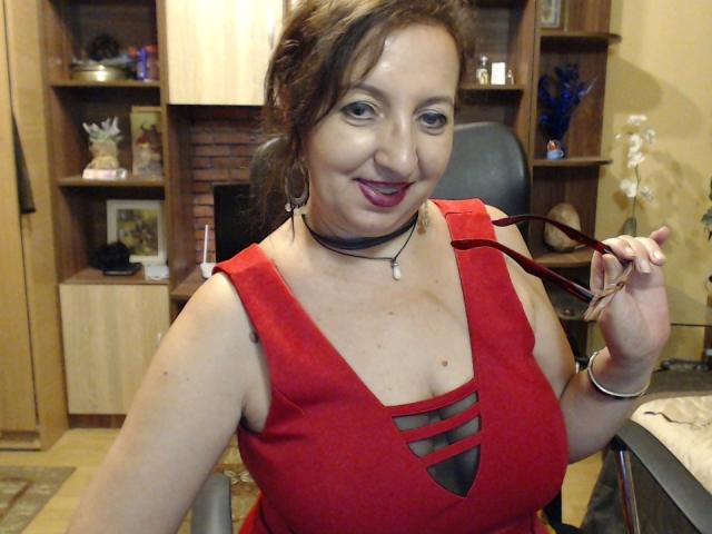 LadyEvelynn, con cabello castaño, vestido rojo y escote, sosteniendo unas gafas rojas, sonriendo, usando un collar negro y una pulsera, en una habitación con estanterías de madera y una chimenea de ladrillo..