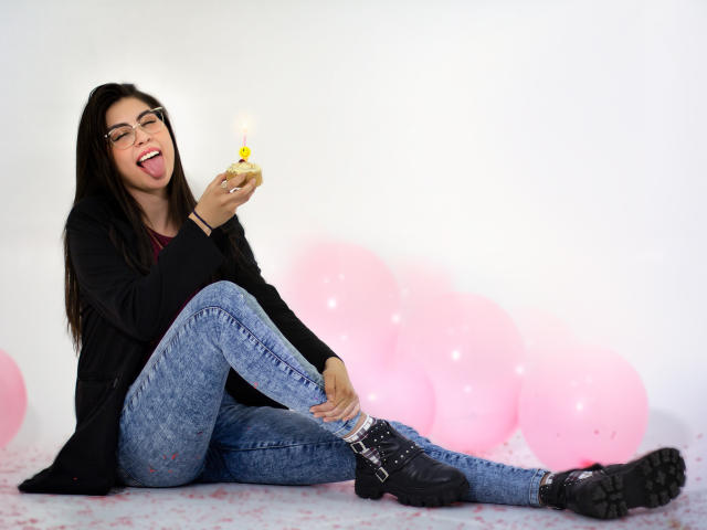 IsaTimeSweet, con lunghi capelli neri, occhiali e una giacca nera, seduta a terra, con la lingua fuori, che tiene una candela accesa, indossa jeans blu e stivali neri, circondata da palloncini rosa..