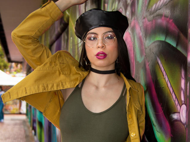 IsaTimeSweet con gli occhiali, una collana nera, una maglietta verde, una giacca gialla, un cappello nero, rossetto rosa, braccio alzato, in piedi di fronte a un muro con graffiti colorati, capelli neri lunghi, seno di media grandezza, che guarda la fotocamera..