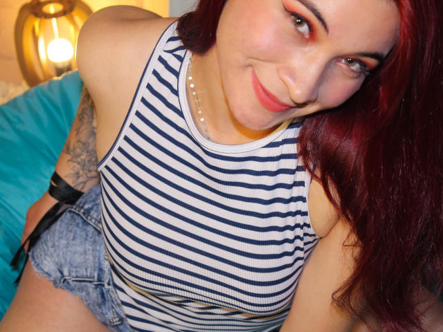 TaniaCespedes with red hair, smiling, wearing a striped tank top, pearl necklace, and denim shorts, showing her tits, tattooed arm, and black wristband.
