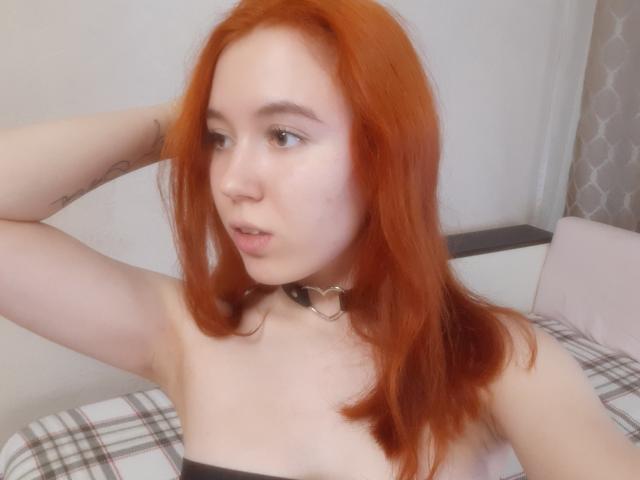 AnikaGlow has red hair, a pale skin, and wears a black strap top with a silver heart choker. She's looking to the side with her arm raised.