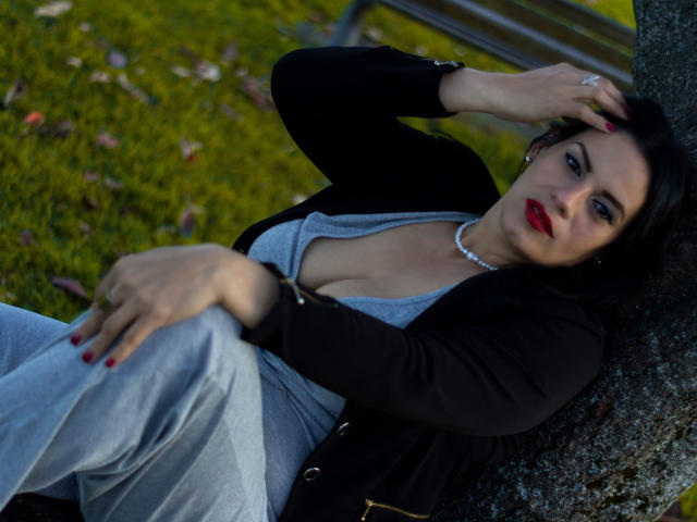LizaRussell, with red lipstick and pearl necklace, leans against a tree, wearing a black jacket and gray dress, showing her tits in a deep cleavage, with red nails.