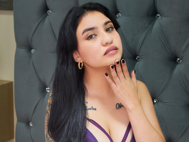 Selena: Angeli con capelli neri, orecchini a cerchio dorati, smalto nero per unghie, lingerie viola, tatuaggi sul petto, mano al collo, che guarda la fotocamera, seduta su una testiera grigia trapuntata..