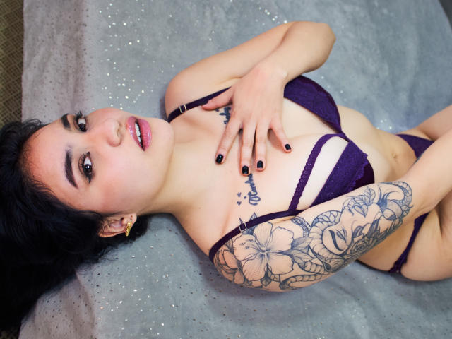 SelenaAngels deitada em uma superfície brilhante, vestindo um sutiã e calcinha de renda roxa, mostrando o braço tatuado com flores, esmalte de unha preto e brincos de ouro, olhando para a câmera com a boca aberta..