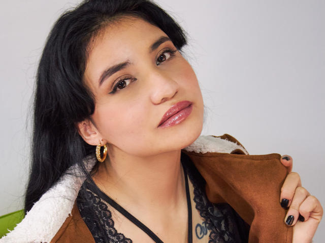 Selena, com lábios rosa brilhantes, delineador preto, brincos de argola dourados, blusa de renda preta, jaqueta marrom e esmalte preto, segurando a gola da jaqueta. Ela tem cabelo escuro e uma tatuagem..