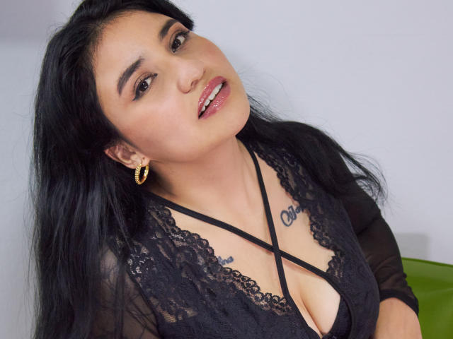 Selena, met lang zwart haar, draagt een zwarte kanten top, gouden oorbellen en een tatoeage boven haar borsten. Ze leunt iets achterover en kijkt met een subtiele glimlach naar de camera..