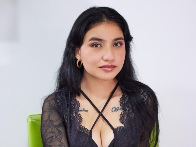 Selena, capelli lunghi e neri, indossa una camicetta di pizzo nero che mette in mostra il seno, ha tatuaggi sul petto, orecchini a cerchio dorati e dietro di lei c'è una sedia verde..
