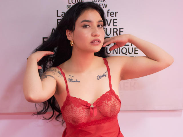 Selena, con un top di pizzo rosso, mostra il seno con tatuaggi sul petto, capelli neri, orecchini dorati e un leggero sorriso, con le mani vicino alla testa..