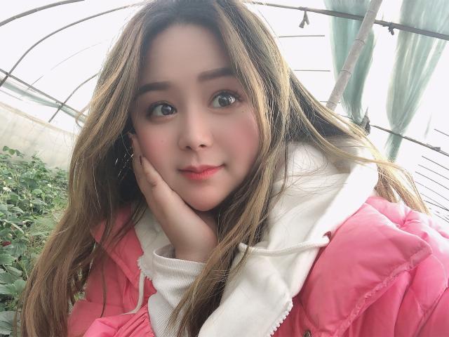 TinaFany, con cabello castaño largo, una chaqueta rosa, una sudadera blanca y una mano en la mejilla, tiene ojos verdes grandes, labios rosados y un rostro lindo, mirando ligeramente hacia la derecha..