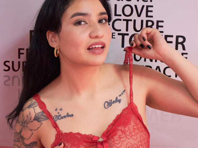 Selena: Angeli con capelli scuri, lingerie di pizzo rossa, orecchini a cerchio dorati, smalto per unghie nero, tatuaggi con i nomi "Elena" e "Cher" sul petto, che si sistema una bretella, sorridente, seno di media grandezza, sfondo rosa tenue..