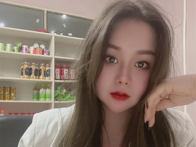 TinaFany, con cabello largo y castaño, lápiz labial rojo y maquillaje rosa, apoyada en su mano, vestida con una camisa blanca, mirando a la cámara con la boca ligeramente abierta..