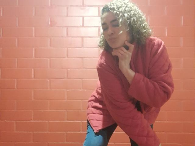 SolmaryHot with curly hair, wearing a pink quilted jacket and blue jeans, leaning forward with a hand on their neck, smiling slightly, in a brightly lit, red-brick room.