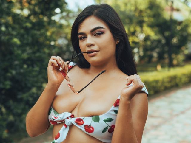 CelesteBelmont mit dunklem Haar, die einen weißen Bikini-Oberteil mit Kirschmuster trägt, eine Sonnenbrille im Mund hält, ihre großen Brüste zeigt und selbstbewusst im Freien steht..