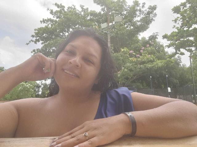 Adi69 with wet brown hair, smiling, wearing a purple top, exposing her bare shoulders and upper chest, has a ring on her right hand, and a bracelet on her left.