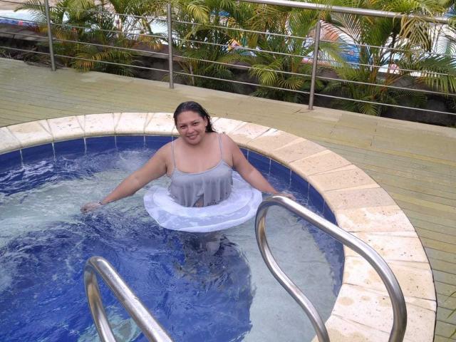 Adi69 con una canottiera trasparente bagnata, che mostra piccoli seni, in una piscina blu rotonda, che tiene un cerchio gonfiabile bianco, sorridendo, in piedi vicino alla scala metallica della piscina..