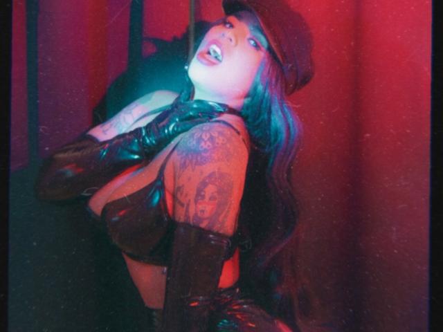 Chili's got her tongue out, face glowing white, tits pushed up, leather gloves, hat, tattooed arm, red and blue light.
