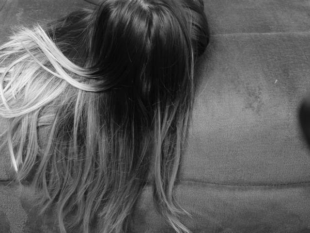 SweetAnaFlowers' head with long, straight, blonde-to-brown ombre hair, laying on a grey couch, visible from the back. Her hair covers her face, looking relaxed.