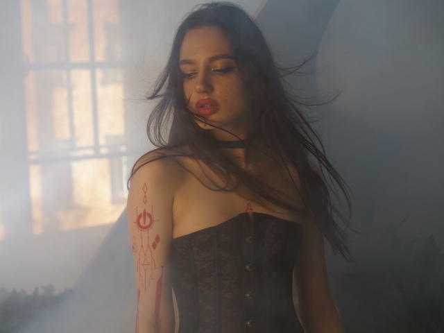 Deviline tem cabelos pretos longos, uma tatuagem vermelha no braço direito, está vestindo um corpete preto e tem lábios vermelhos cheios. A luz suave e a névoa criam uma atmosfera sensual e misteriosa..