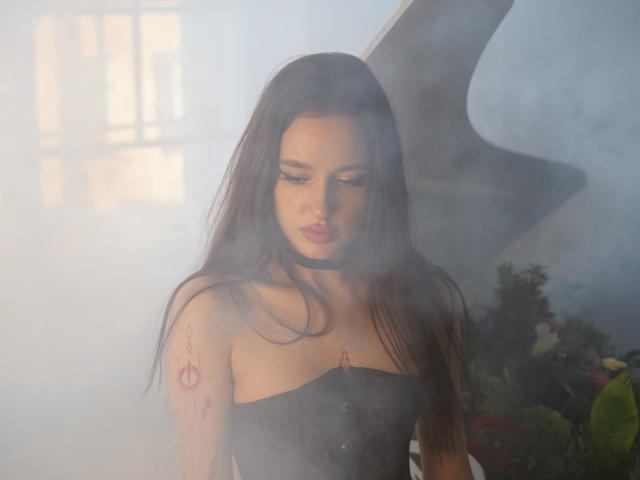 Deviline, com longos cabelos castanhos, vestindo uma blusa preta sem alças, olhando para baixo com um leve semblante de descontentamento. Ela tem uma tatuagem rosa no braço direito e um colar preto..