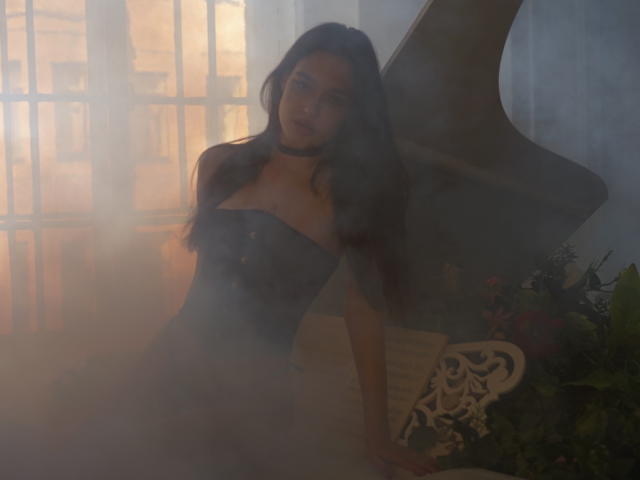 Uma mulher com cabelo escuro, usando um colar preto e um vestido sem alças, sentada em uma luz fraca e nebulosa. Seus seios estão visíveis, com os braços apoiados em uma cadeira branca ornamentada..