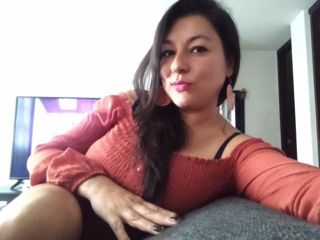SakotoDika with dark hair, pink lipstick, and red off-shoulder top, showing black bra strap, sitting with hand on gray chair, slightly smiling. Boobs visible under top.