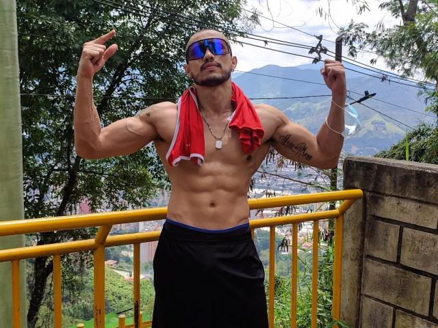 DimitriRivas, sin camisa, musculoso, bronceado, toalla roja alrededor del cuello, pantalones cortos negros, visera azul, tatuajes, señalando con ambas manos, de pie sobre una barandilla amarilla, montañas en el fondo..