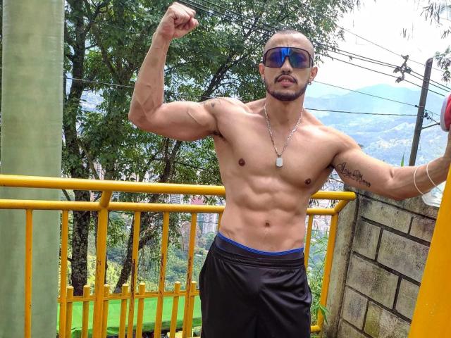 Muscular DimitriRivas with a shaved head and beard, flexing his right arm, wearing blue sunglasses, black shorts, and a dog tag necklace, standing outside with yellow railings.