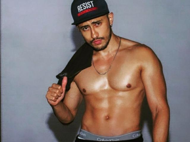 DimitriRivas, sin camisa, musculoso, con piel bronceada, usando un gorro negro y ropa interior Calvin Klein, sosteniendo una camisa negra sobre su hombro derecho, haciendo un gesto de pulgar hacia arriba, con expresión seria..