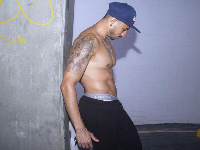 Shirtless DimitriRivas with tattoos on his right arm, wearing a blue cap and black Calvin Klein pants, leaning against a concrete wall, showing a toned, muscular, brown body.