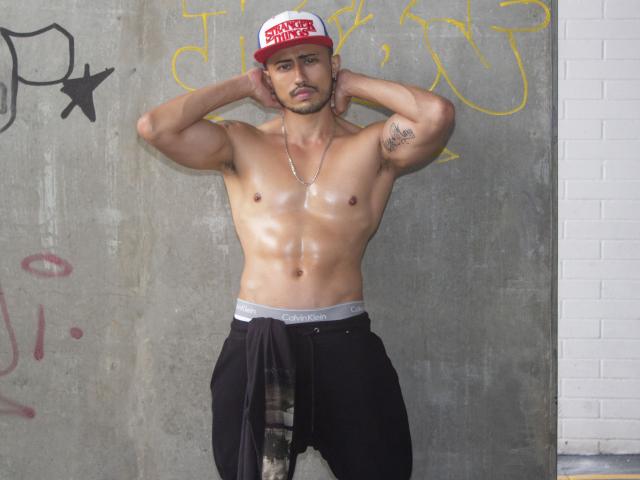 Shiny, muscular DimitriRivas with a tattoo on his right arm, wearing a red and white cap, black pants, and a silver necklace, standing with hands behind head, showing his ass.