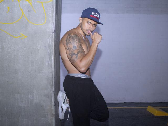 DimitriRivas, sin camisa, con un brazo musculoso y tatuado, pantalones negros y una gorra azul, de pie en un espacio de hormigón, con los puños levantados, zapatillas blancas, mirando a la cámara..