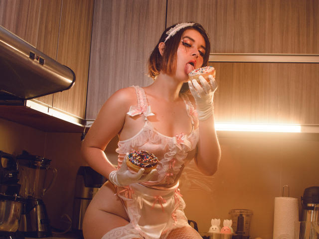 MaraMaison con i seni scoperti, che indossa un grembiule rosa con volant, guanti bianchi e una fascia per capelli, mentre mangia una ciambella, tenendone un'altra in mano, in una cucina, con una macchina da caffè sullo sfondo..