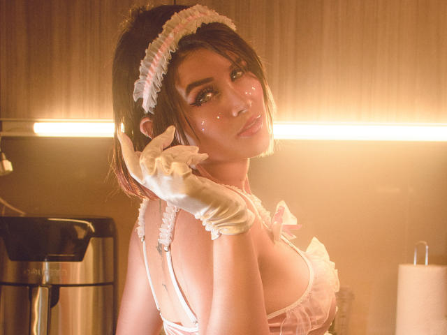 MaraMaison, com um rosto brilhante, uma tiara branca com babados, um sutiã rosa, luvas brancas, segurando o cabelo, olhando para trás, em uma luz quente, mostrando seus seios e uma expressão fofa e sexy..