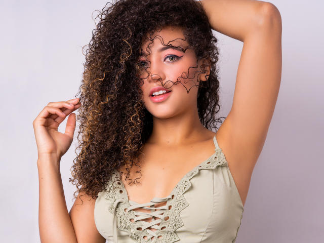 AlyssaBelle, com cabelo castanho encaracolado, blusa verde com cadarços, braço levantado e linhas de maquiagem preta onduladas no rosto, sorrindo para a câmera. Ela está mostrando seus seios..