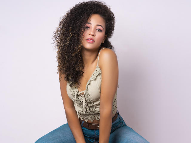 AlyssaBelle, com cabelos castanhos cacheados, pele bronzeada, vestindo uma blusa bege com babados e jeans azuis, sentada com uma leve inclinação para frente, mostrando seus pequenos seios e um cinto marrom..