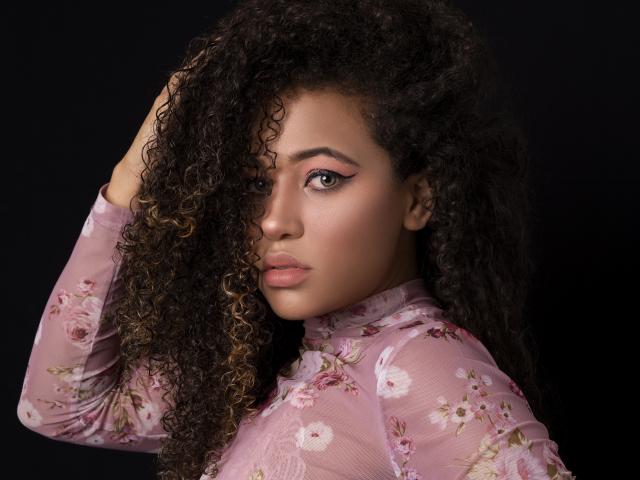 AlyssaBelle, com cabelos castanhos cacheados, vestindo uma blusa rosa transparente com estampas florais, tem a mão no cabelo, olhando com olhos verdes, sombra rosa e uma expressão séria..
