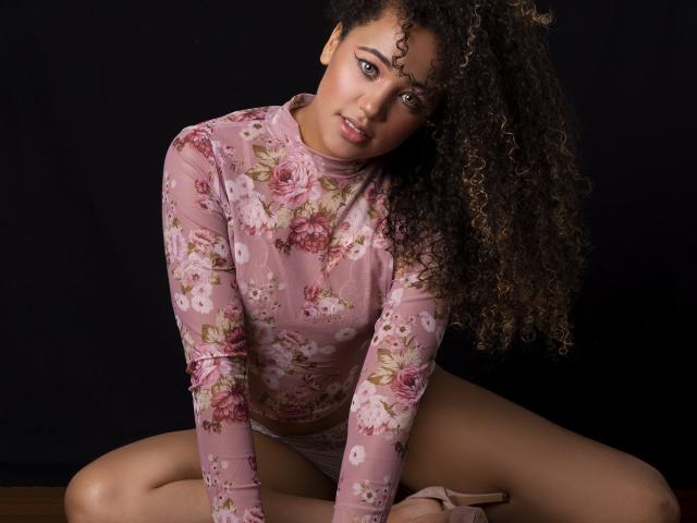 AlyssaBelle with curly brown hair, wearing a pink floral long-sleeve top, sitting with legs apart, showing her pussy, looking at the camera with a slight smile.