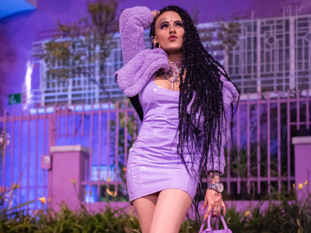 MollyWaxx with long black braids, purple shiny latex dress, fluffy purple sleeves, standing, hand in hair, looking up, holding purple bag, purple light, purple background, sexy, confident.