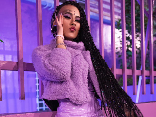 MollyWaxx mit langen schwarzen Zöpfen, die einen violetten, flauschigen Jacke und glänzende pinke Hosen trägt, mit violetten Lichtern und violetten Wandstangen, Hand in der Nähe des Ohrs, leicht schmollende Lippen, Ring am Handgelenk..