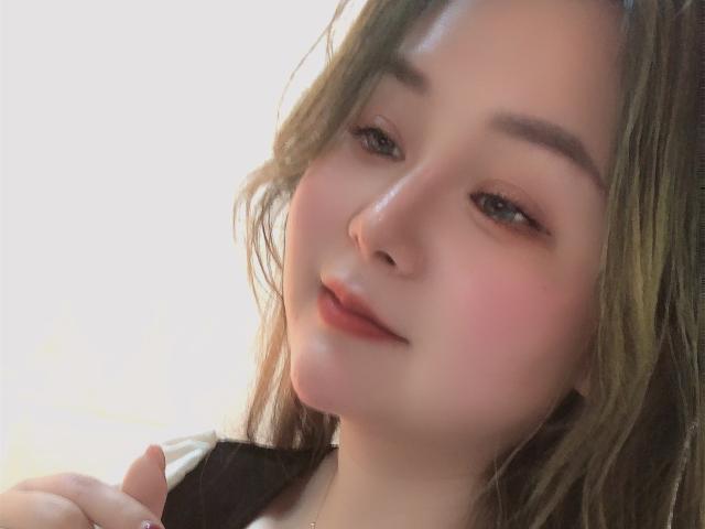 TinaFany tiene una piel clara, cabello ondulado de color verde-marrón y una sonrisa sutil. Se pueden ver sus mejillas rosadas, sus cejas marrones claras y sus labios rojos. Su mano toca su camisa negra..